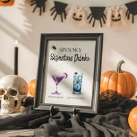 A framed Halloween-themed sign reading 'Spooky Signature Drinks' with two illustrated cocktails, displayed on a table surrounded by pumpkins, a skull, black fabric, and a candle in a decorated indoor setting.
