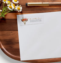 White envelope with floral design and text on a wooden surface with flowers and a pen.