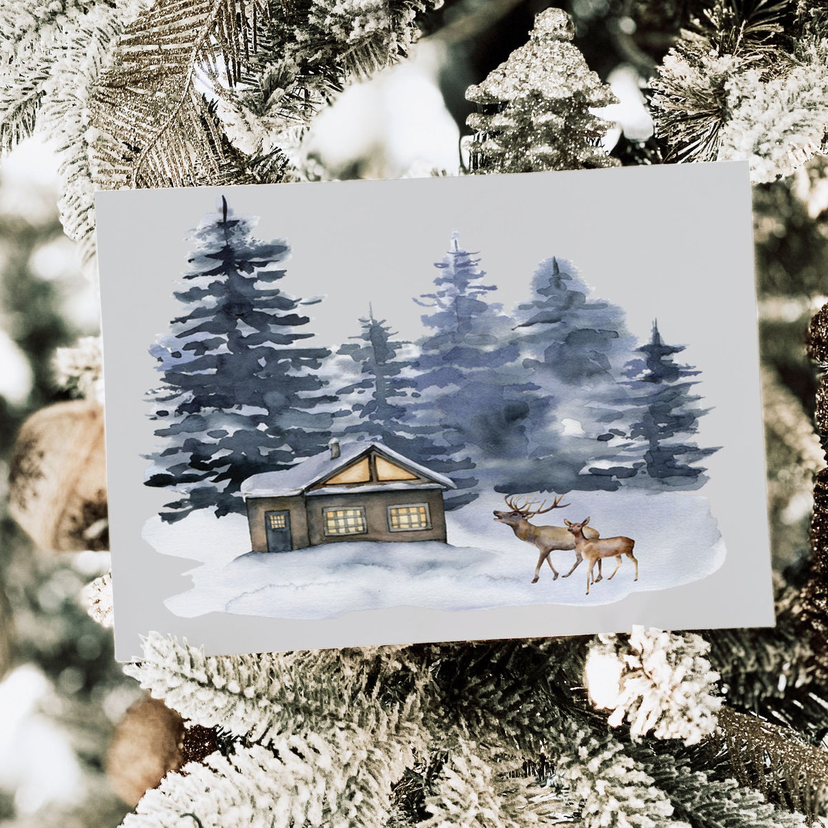 A rectangular art print or card featuring a winter forest scene with a small cabin, snow, and two deer, displayed among frosted Christmas tree branches.