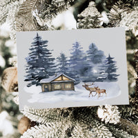 A rectangular art print or card featuring a winter forest scene with a small cabin, snow, and two deer, displayed among frosted Christmas tree branches.