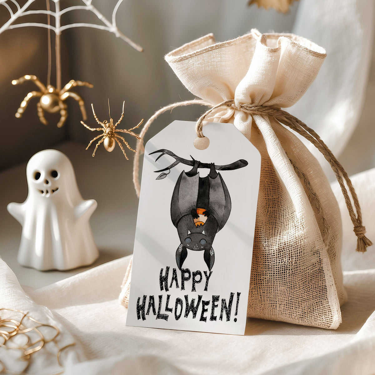 A small beige burlap drawstring pouch tied with twine and a white hanging tag printed with a bat illustration and the words 'HAPPY HALLOWEEN!', styled on draped fabric with Halloween props including a ghost figurine, spiders, and a web decoration.