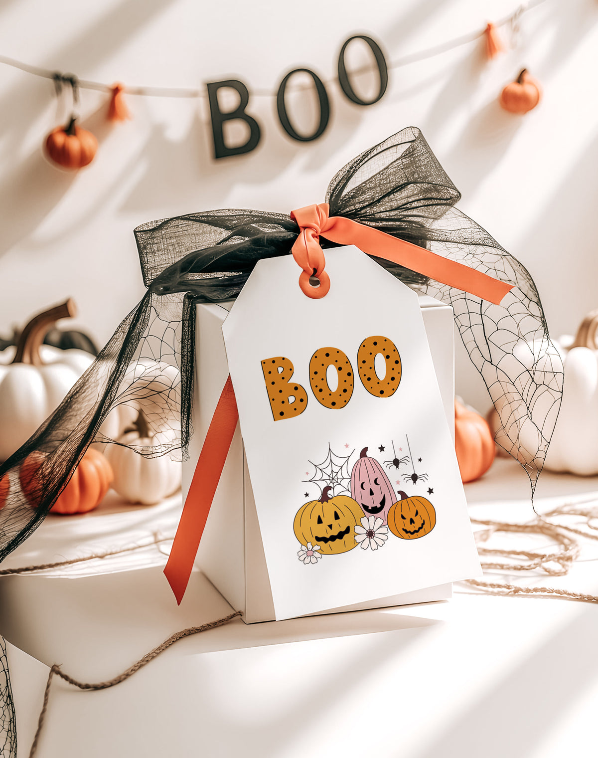 A white Halloween gift tag with 'BOO' in cookie letters and jack-o'-lantern illustrations, tied with a black tulle bow and orange ribbon, styled on a gift box with pumpkins and a 'BOO' banner in the background.