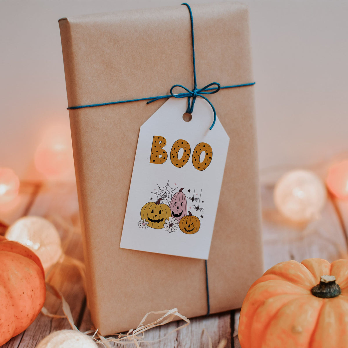 A wrapped gift box in brown kraft paper with a blue string and a white 'BOO' tag, styled with small pumpkins and warm string lights on a wooden surface.