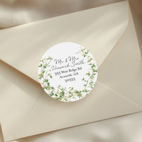 A cream-colored envelope sealed with a round white sticker label featuring a green leaf wreath and elegant script text, styled on draped satin fabric with dried pampas grass.