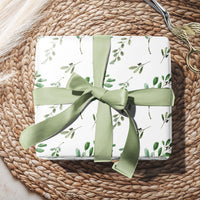 Gift box with green ribbon and floral pattern on a woven surface with eucalyptus leaves.