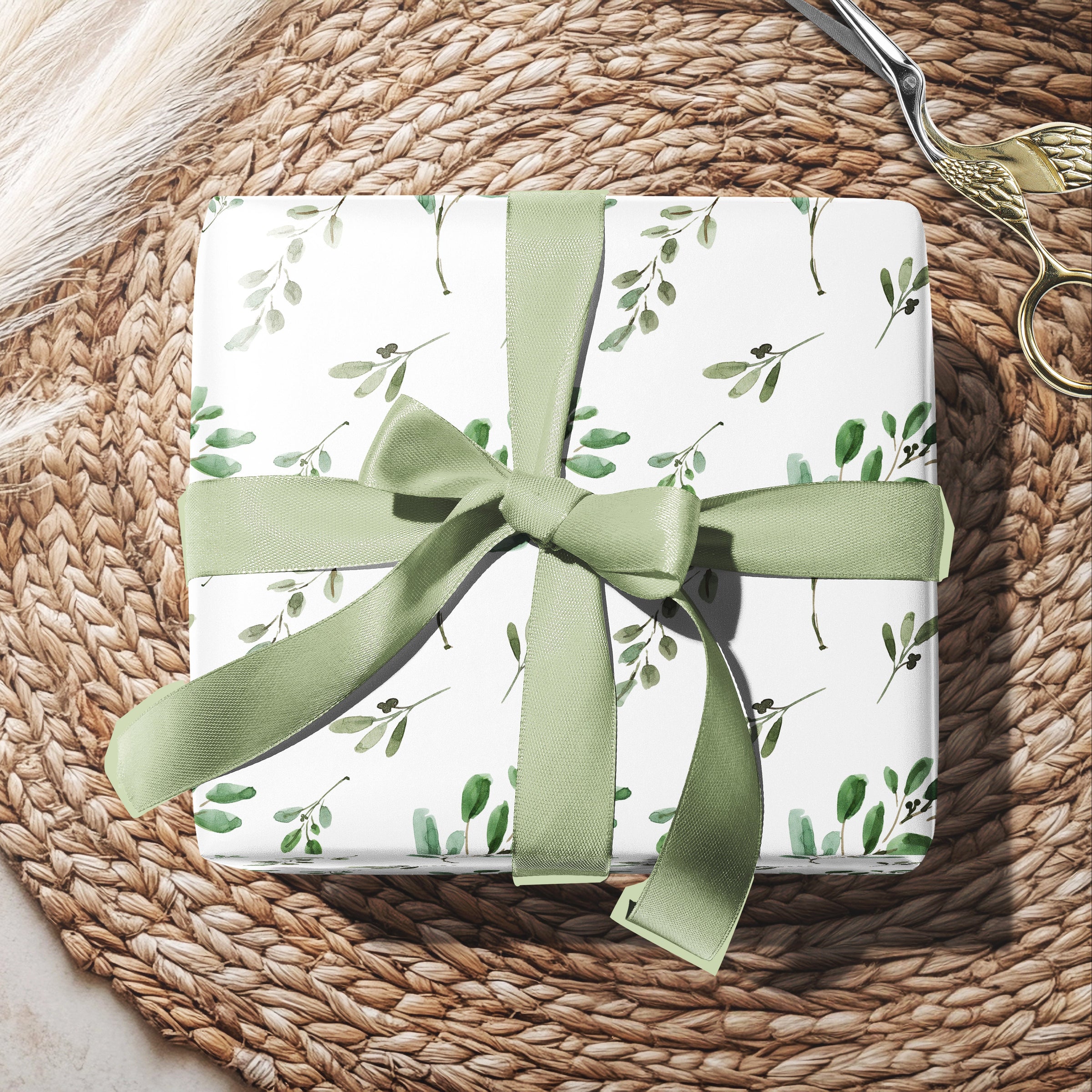Gift box with green ribbon and floral pattern on a woven surface with eucalyptus leaves.