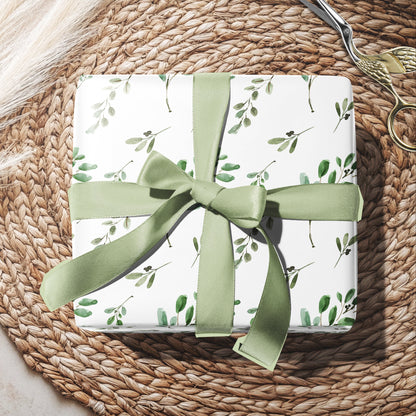 Gift box with green ribbon and floral pattern on a woven surface with eucalyptus leaves.