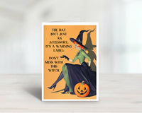 A Halloween greeting card featuring a vintage-style illustration of a witch sitting with a jack-o'-lantern, with the text 'THE HAT ISN'T JUST AN ACCESSORY, IT'S A WARNING LABEL: DON'T MESS WITH THIS WITCH.' The card is shown standing upright on a light surface against a soft white background.