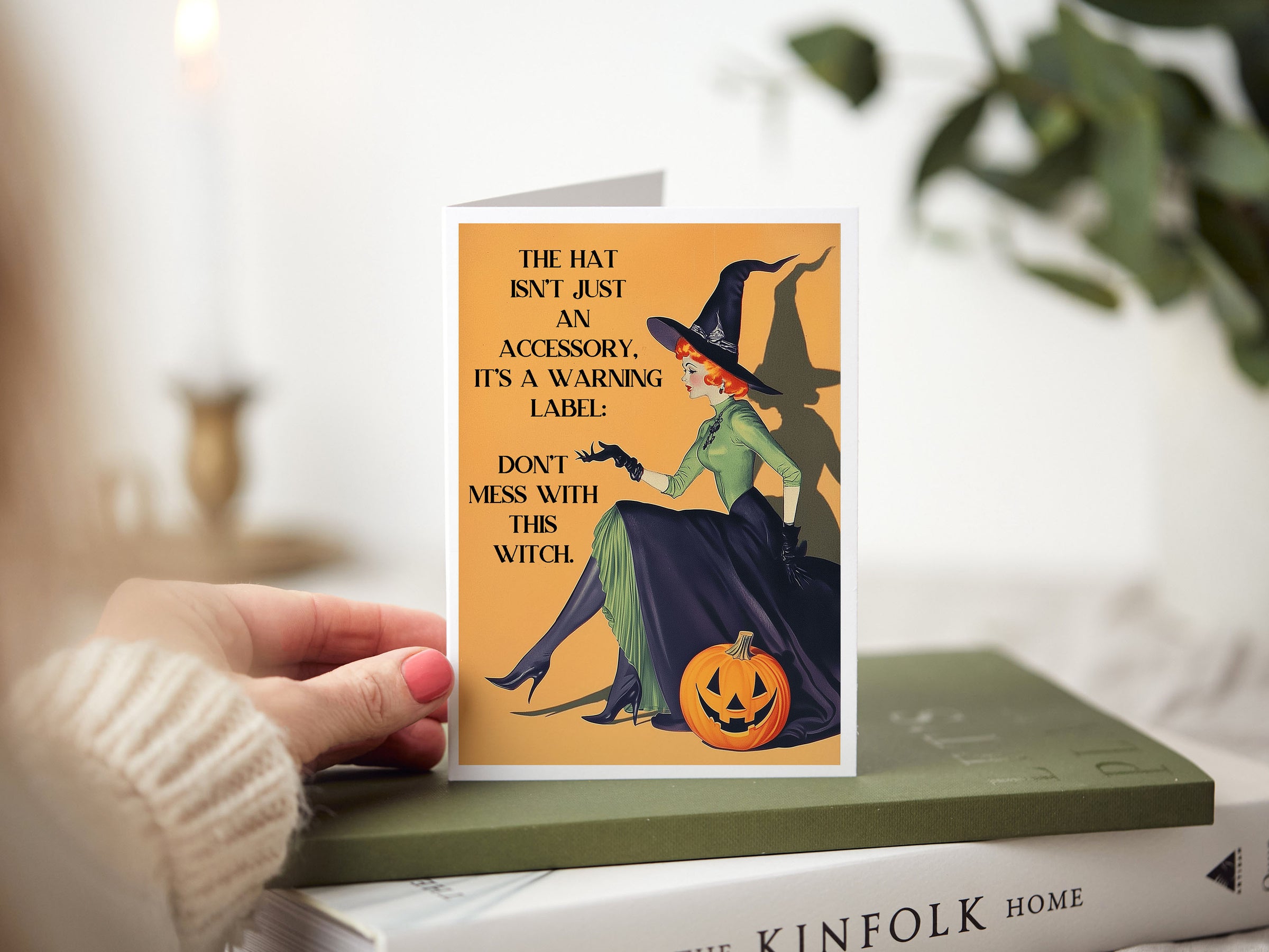 A hand holds a folded greeting card featuring a vintage-style illustration of a witch sitting on a pumpkin, with humorous text, set against a softly blurred indoor background with books and greenery.