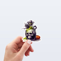 A hand holds a die-cut sticker featuring a skull inside a dark potion bottle with green slime, a spider on top, and a small orange pumpkin beside it, against a light gray background.