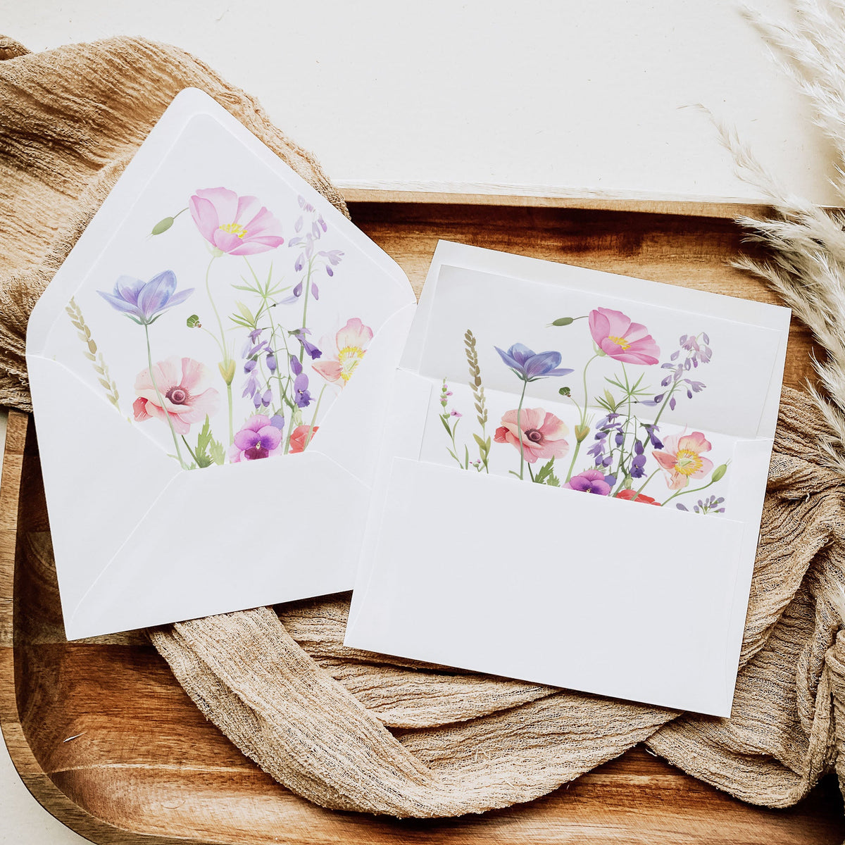 A set of white stationery cards and envelopes featuring a colorful watercolor floral design, styled on a rustic wooden tray with beige fabric.