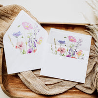 A set of white stationery cards and envelopes featuring a colorful watercolor floral design, styled on a rustic wooden tray with beige fabric.
