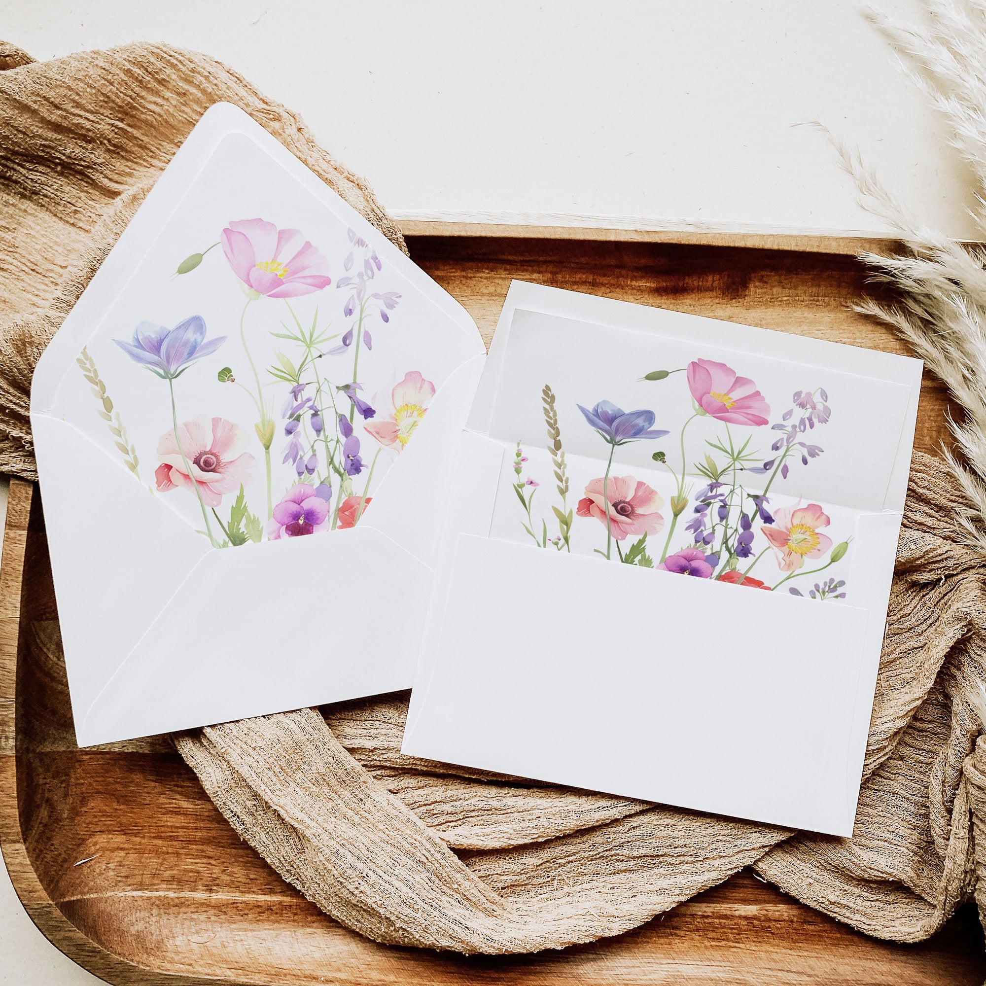 A set of white stationery cards and envelopes featuring a colorful watercolor floral design, styled on a rustic wooden tray with beige fabric.