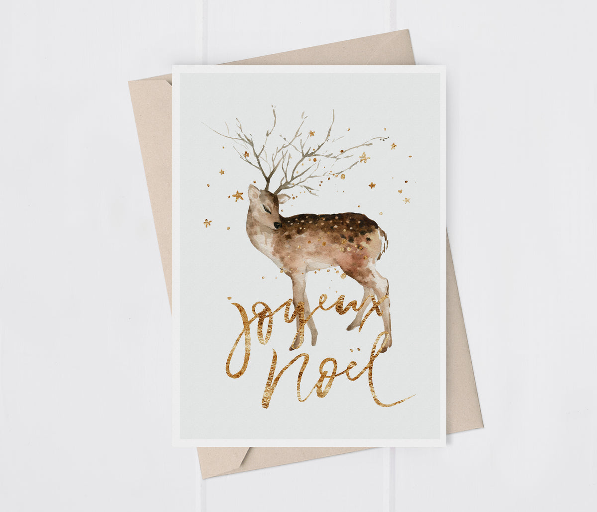 A white greeting card with a watercolor deer illustration, gold script reading 'Joyeux Noel', and small gold stars, shown with a kraft envelope on a white background.