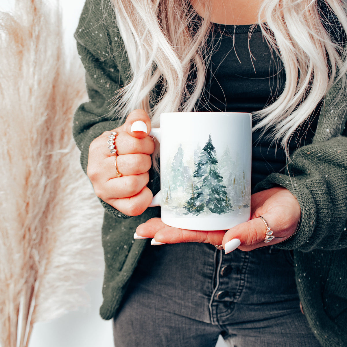 A person holding a white ceramic mug with a winter forest design, wearing a dark green knit cardigan and black top, with blonde hair and rings visible.