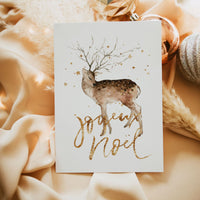A white greeting card featuring a watercolor deer and gold glittery 'Joyeux Noel' script, styled on draped beige satin fabric with decorative ornaments.